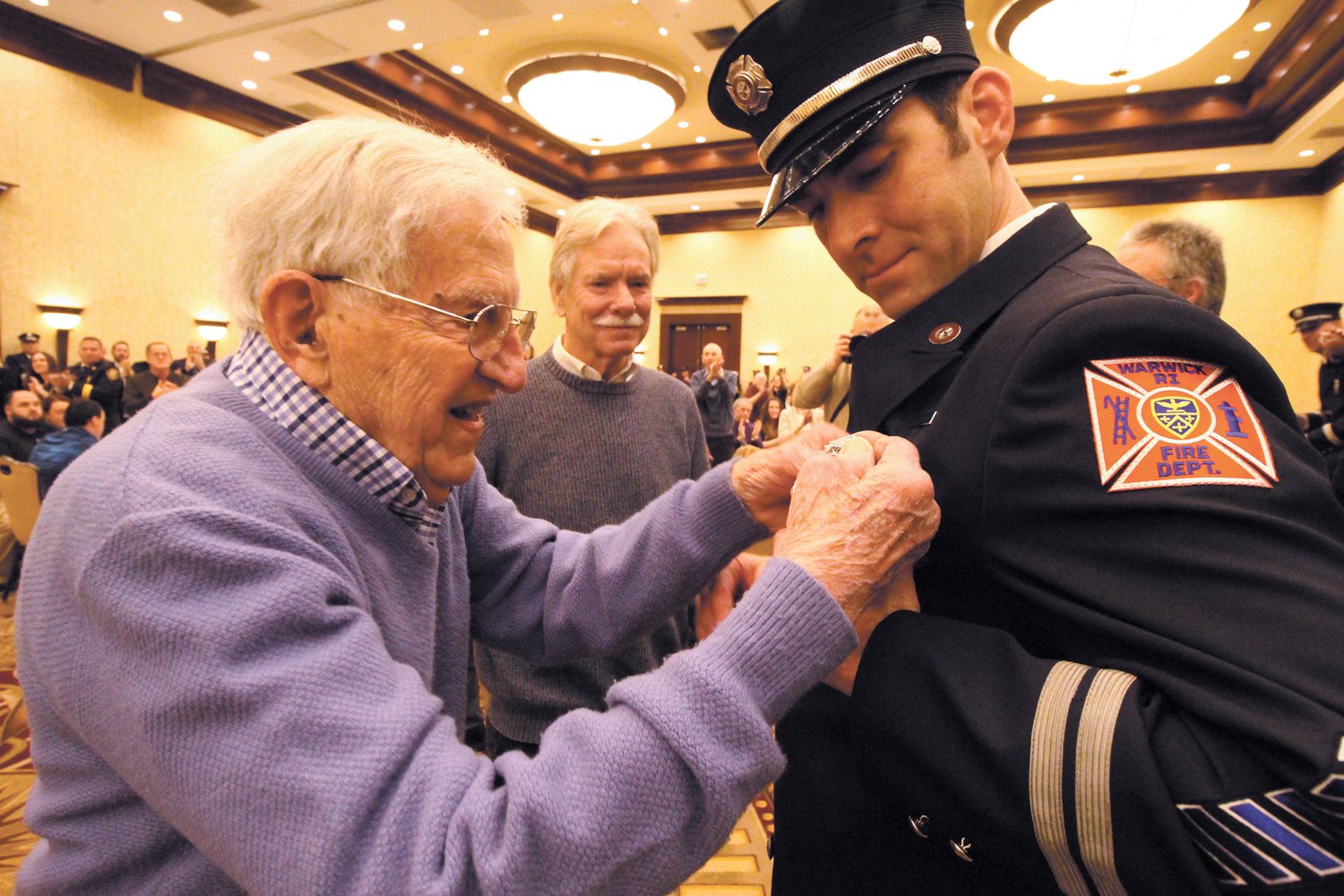 Their service applauded: More than 300 people stood to applaud when WWII veteran Maurice Roland Boulais participated in  the promotional ceremony of 21 firefighters Wednesday night at the Crowne Plaza. Boulais with his son-in law Henry Andrews standing by affixed the badge of Assistant Fire Marshal Ethan Andrews, Henry’s son. (Warwick Beacon photos)